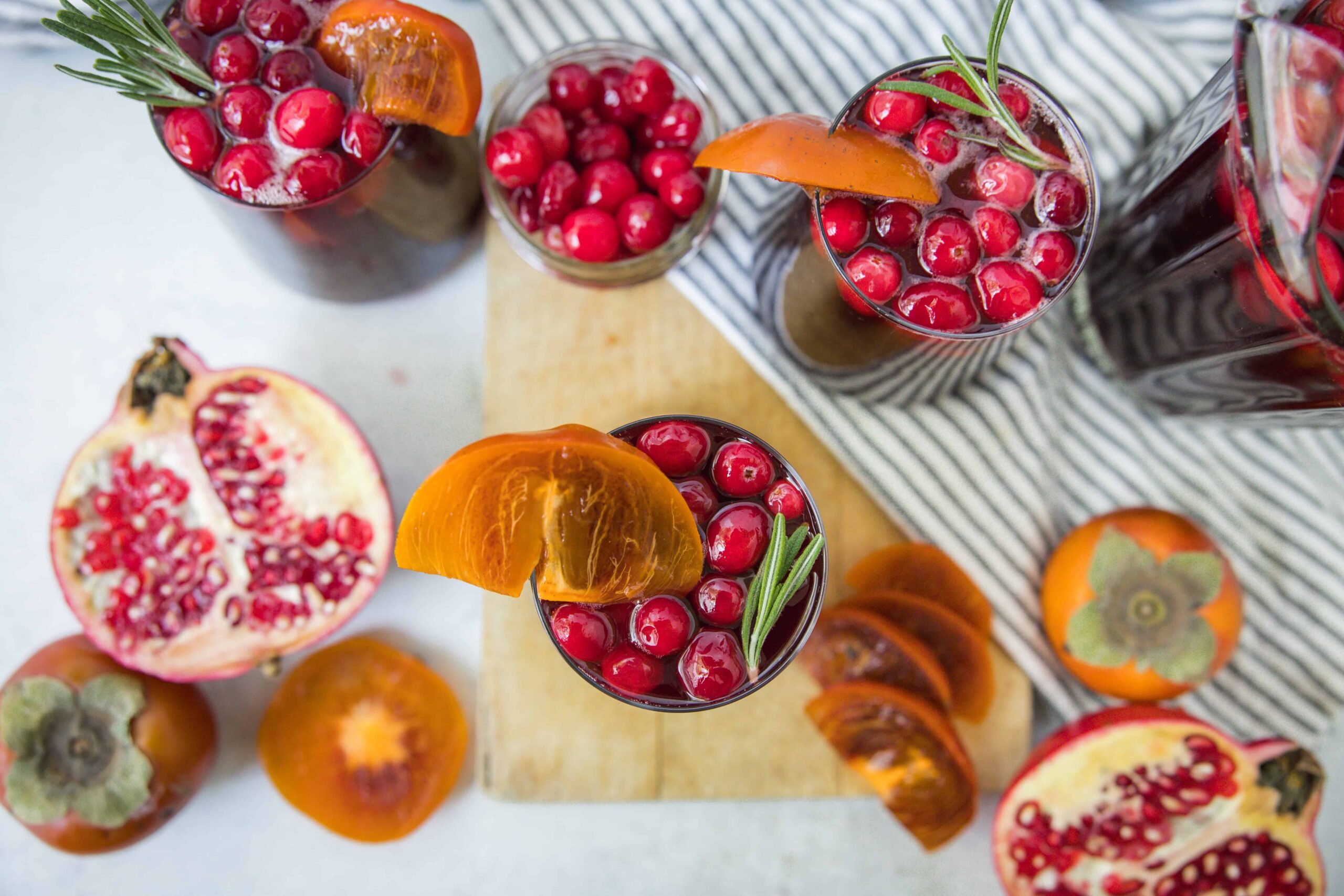 Juicy pomegranate and persimmon fruit drinks garnished with rosemary, served in glasses on wooden board with fresh fruits and striped cloth background, perfect for fall and winterrefreshing beverage.