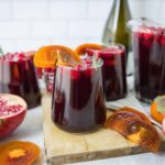 Rich pomegranate juice with orange slices garnished with rosemary, served in glasses, featuring fresh pomegranate and persimmon fruits, ideal for healthy drink recipes and fruit-based beverages.