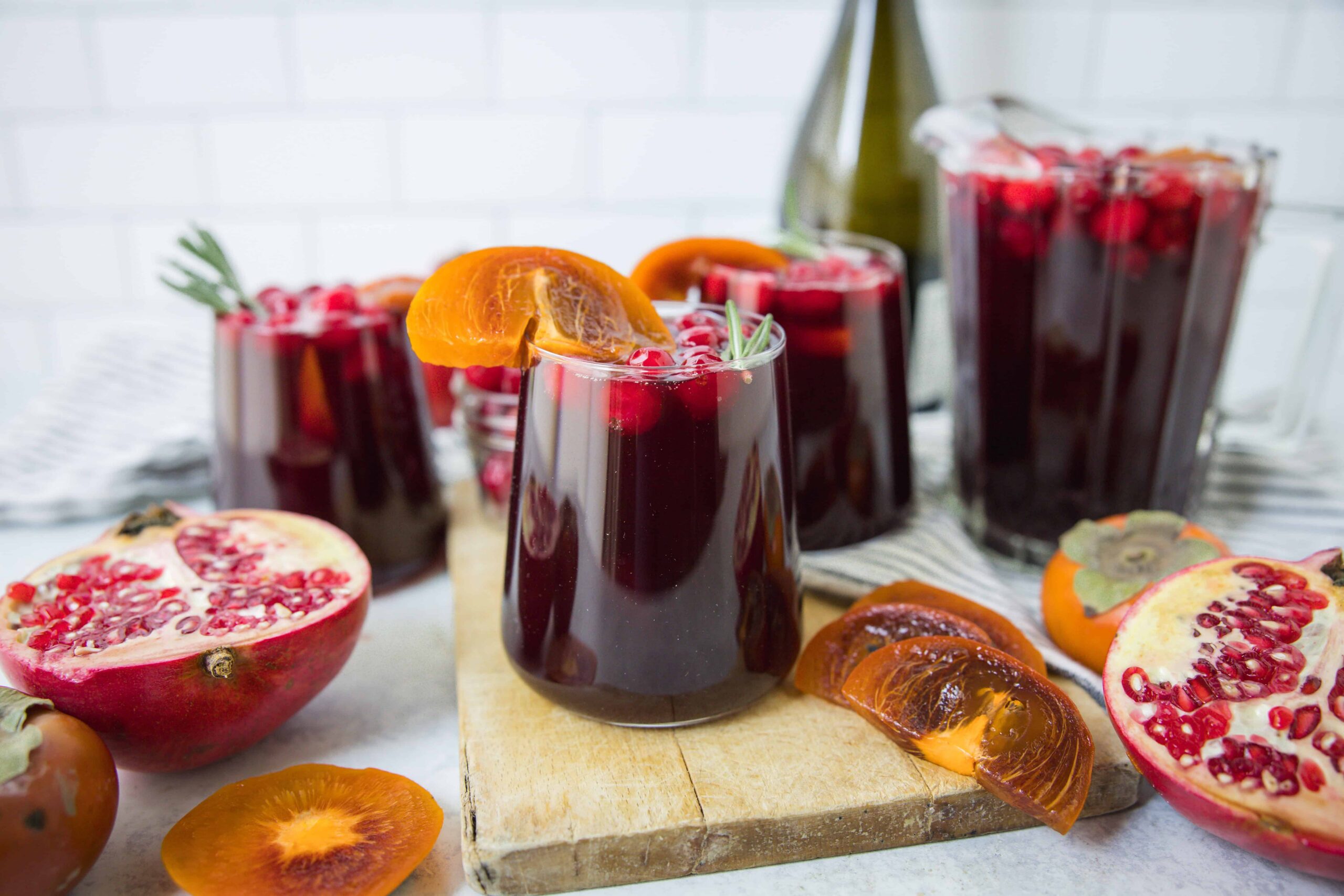 Rich pomegranate juice with orange slices garnished with rosemary, served in glasses, featuring fresh pomegranate and persimmon fruits, ideal for healthy drink recipes and fruit-based beverages.