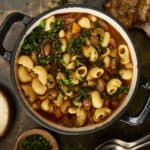 Hearty vegetable and pasta soup in a black pot with chopped herbs, served with bread and grated cheese on a rustic wooden surface, perfect for cozy meals and nutritious comfort food.