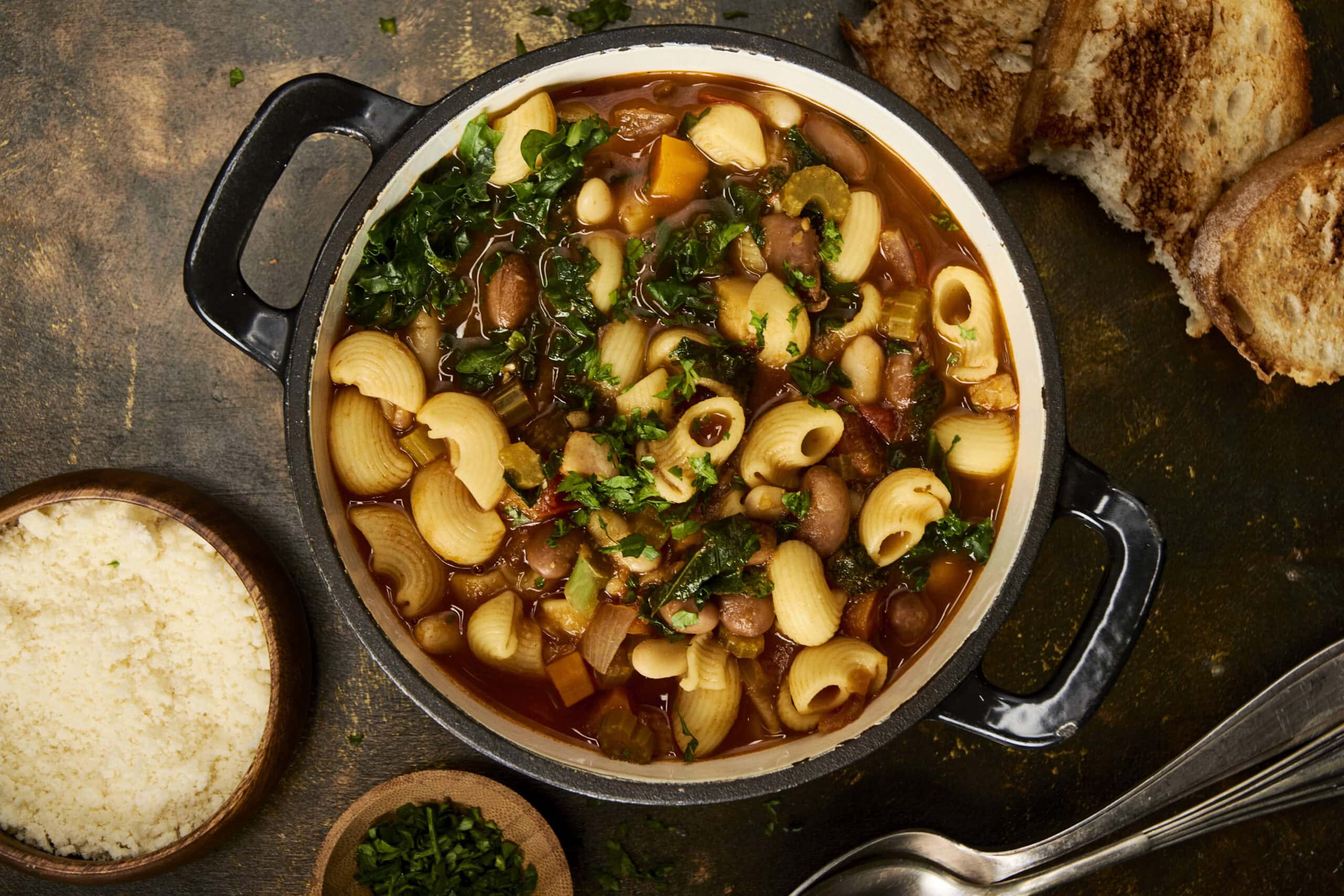 Hearty vegetable and pasta soup in a black pot with chopped herbs, served with bread and grated cheese on a rustic wooden surface, perfect for cozy meals and nutritious comfort food.