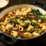 Creamy vegetable pasta soup with kale beans and pasta in a black skillet on dark wooden background.