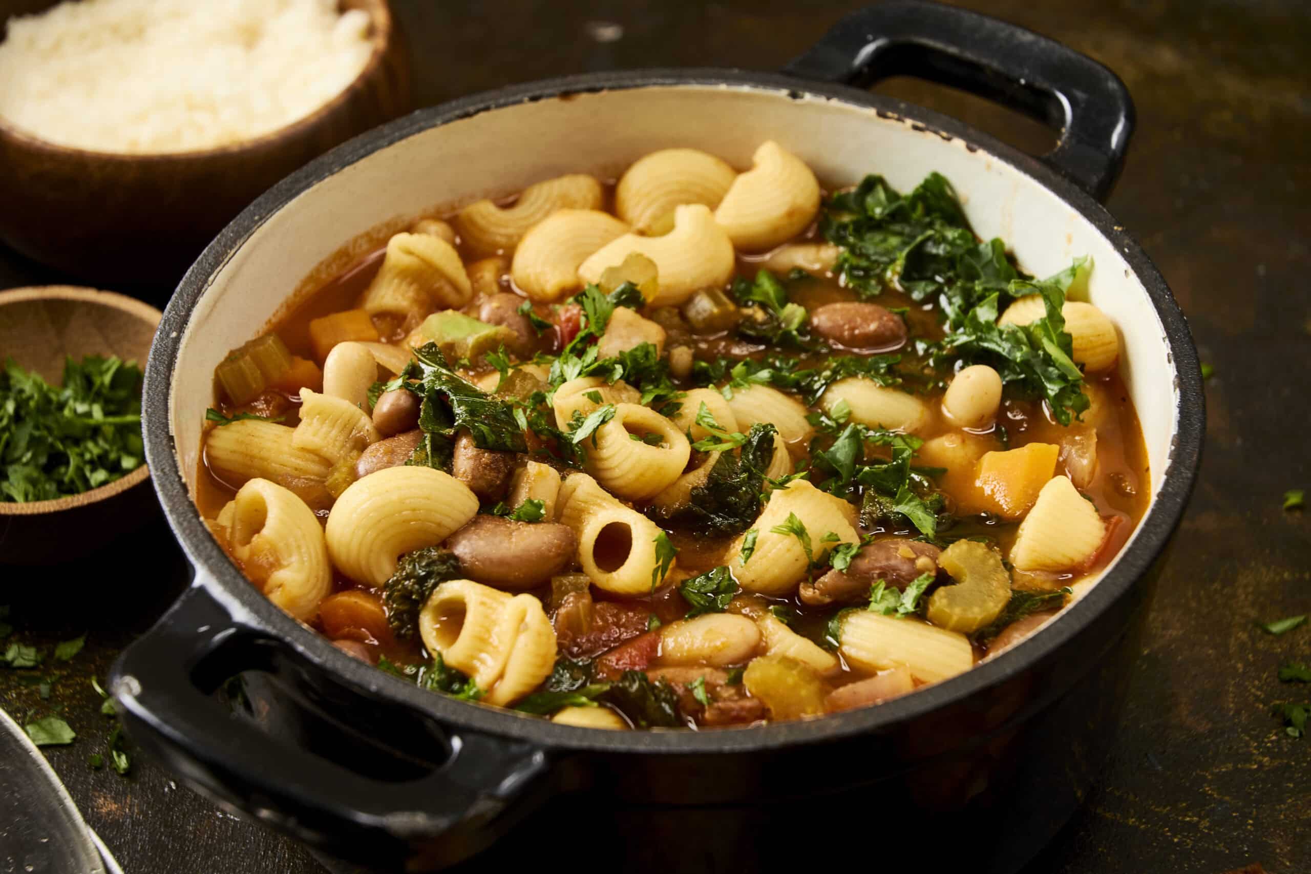 Creamy vegetable pasta soup with kale beans and pasta in a black skillet on dark wooden background.