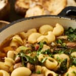 Hearty vegetable and pasta soup in a rustic white pot, topped with fresh herbs and served with toasted bread in the background, perfect for cozy meals and comfort food.