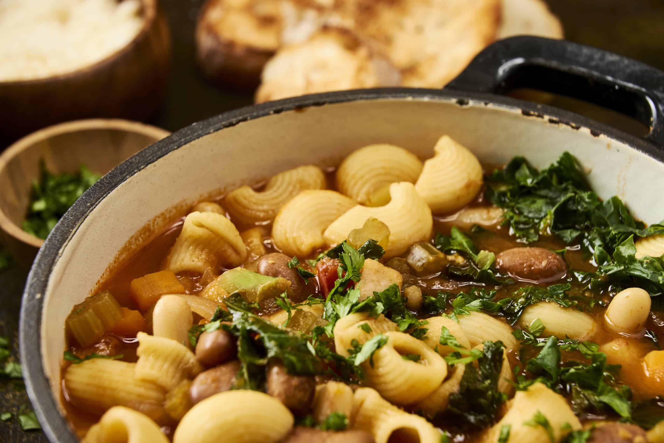 Hearty vegetable and pasta soup in a rustic white pot, topped with fresh herbs and served with toasted bread in the background, perfect for cozy meals and comfort food.