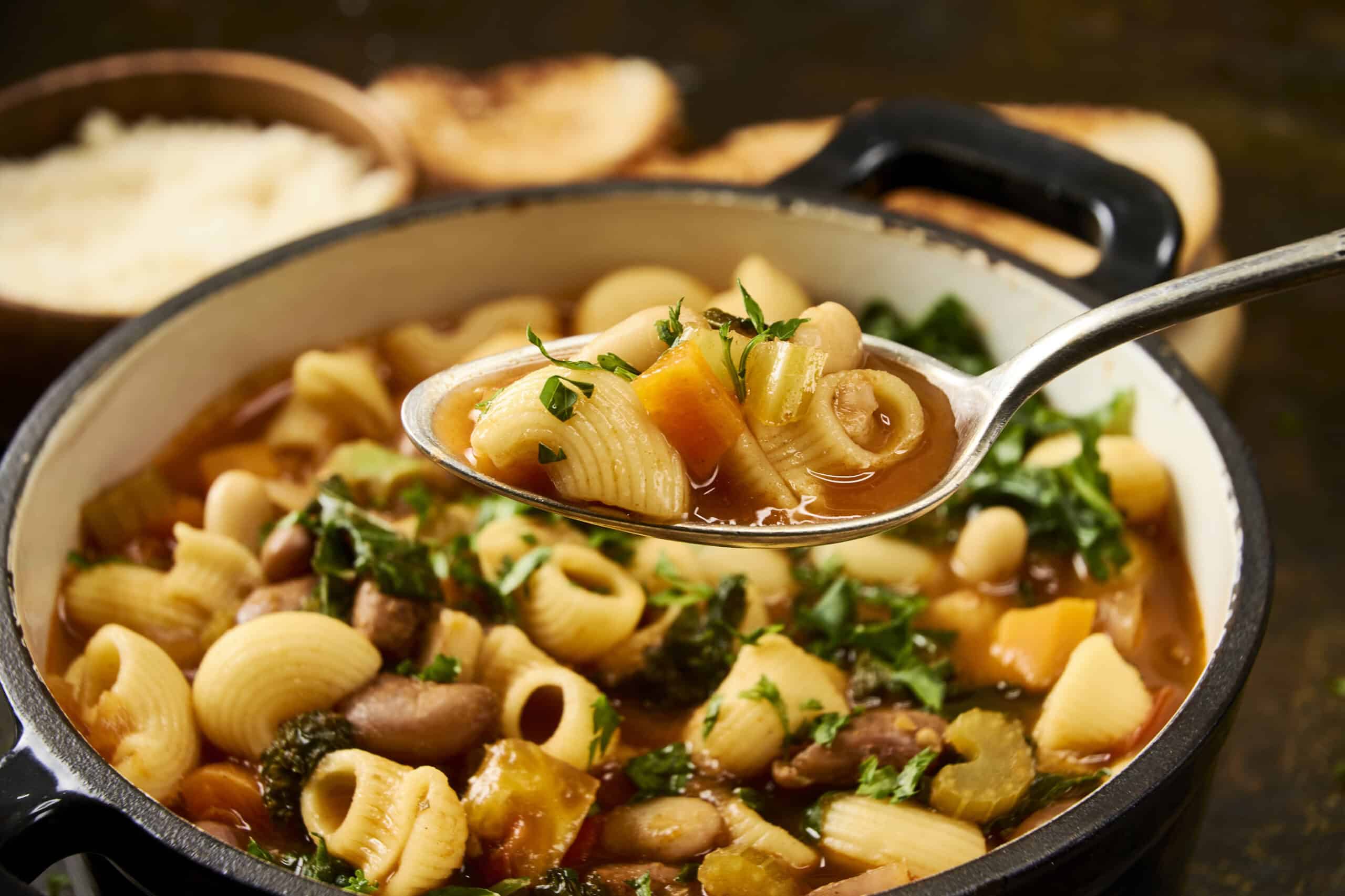 Creamy pasta vegetable soup with herbs in a black pot, ready to serve, fresh vegetables and pasta pieces visible in the hearty meal.