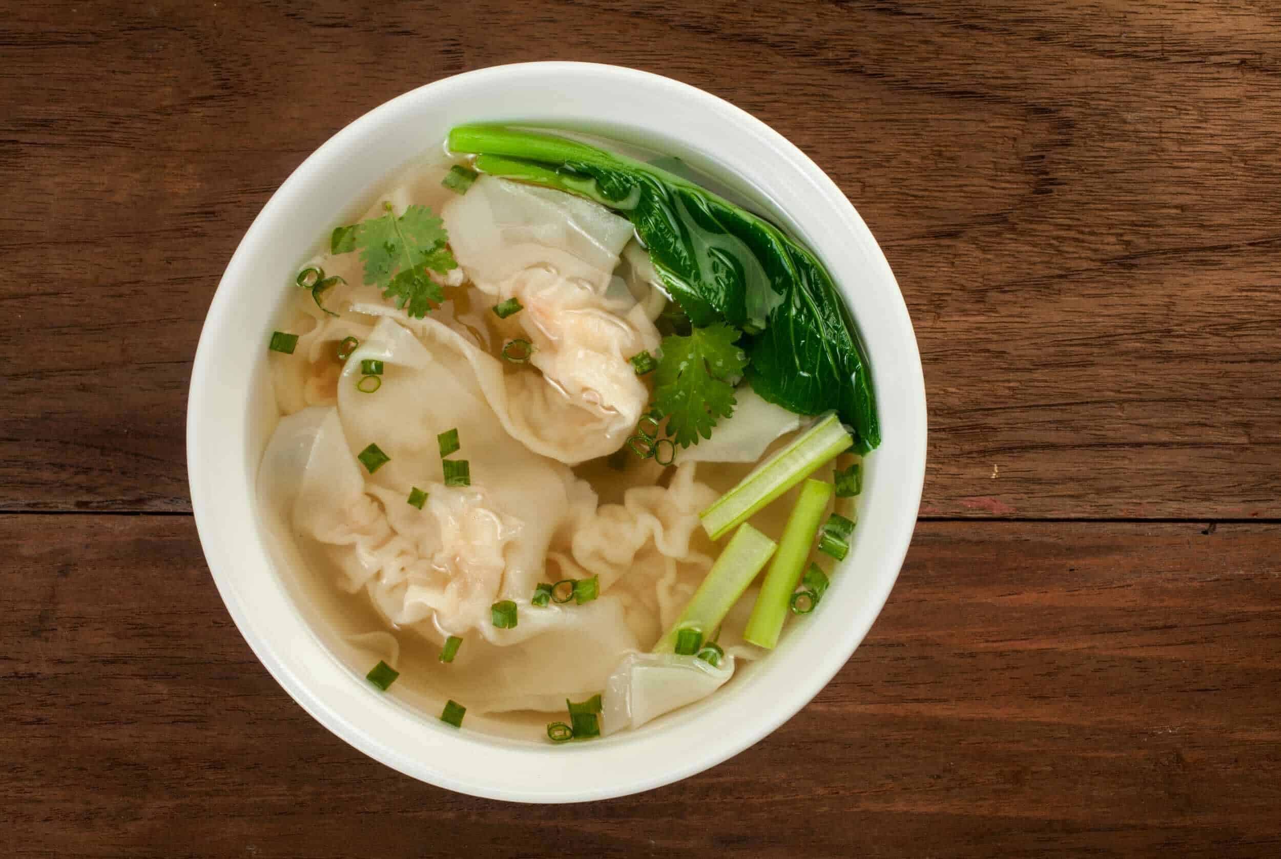 Steamed dumpling soup with green vegetables and fresh herbs in a white bowl on a wooden surface.