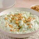 Creamy cucumber salad topped with chopped nuts in a white dotted bowl, with bowls of nuts and creamy dressing in the background, perfect for a fresh, crunchy side dish.
