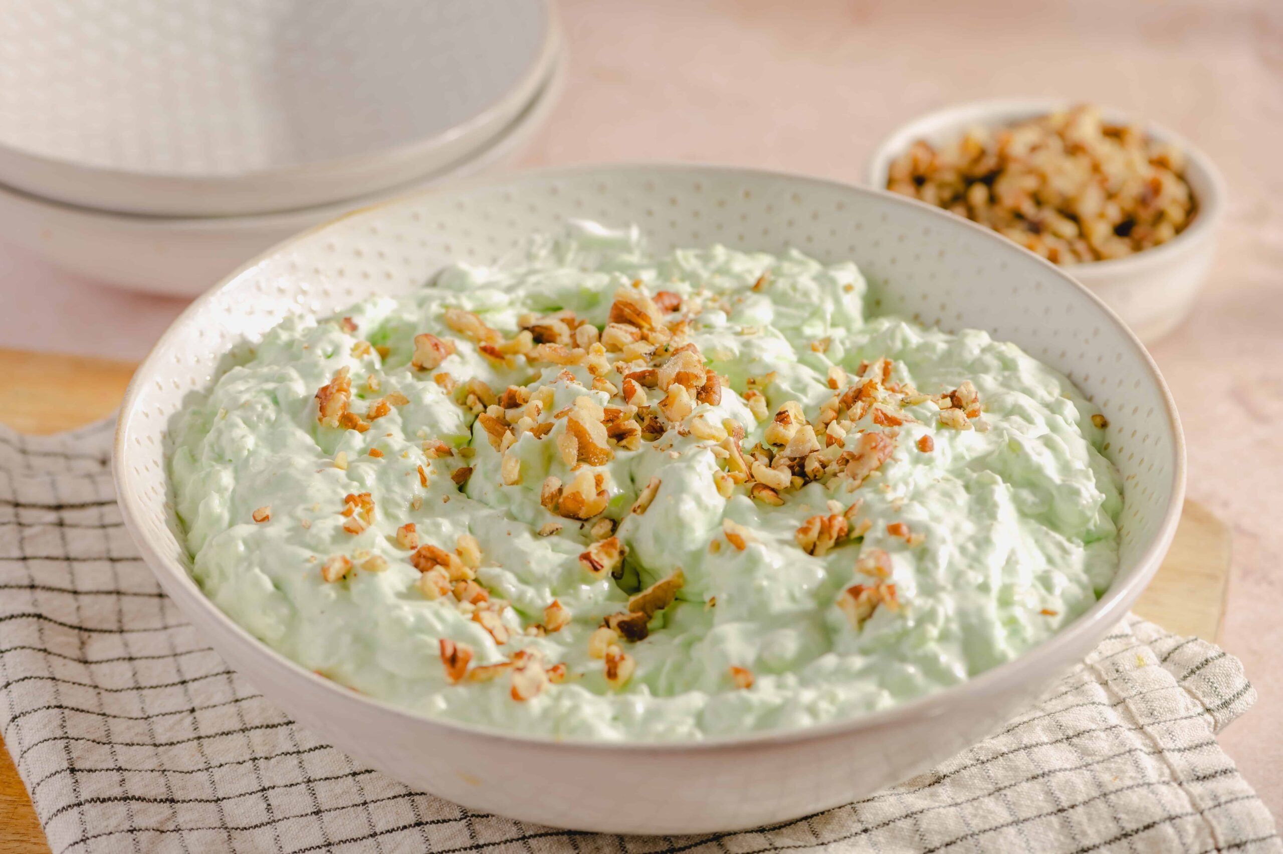 Creamy cucumber salad topped with chopped nuts in a white dotted bowl, with bowls of nuts and creamy dressing in the background, perfect for a fresh, crunchy side dish.