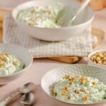 Creamy cucumber salad with chopped nuts, served in textured white bowls on a pink surface with a spoon, featuring crispy croutons or toppings, with additional bowls of salad and chopped nuts in the background.