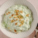 Creamy avocado salad topped with crushed nuts in a white honeycomb-patterned bowl on a light pink surface.