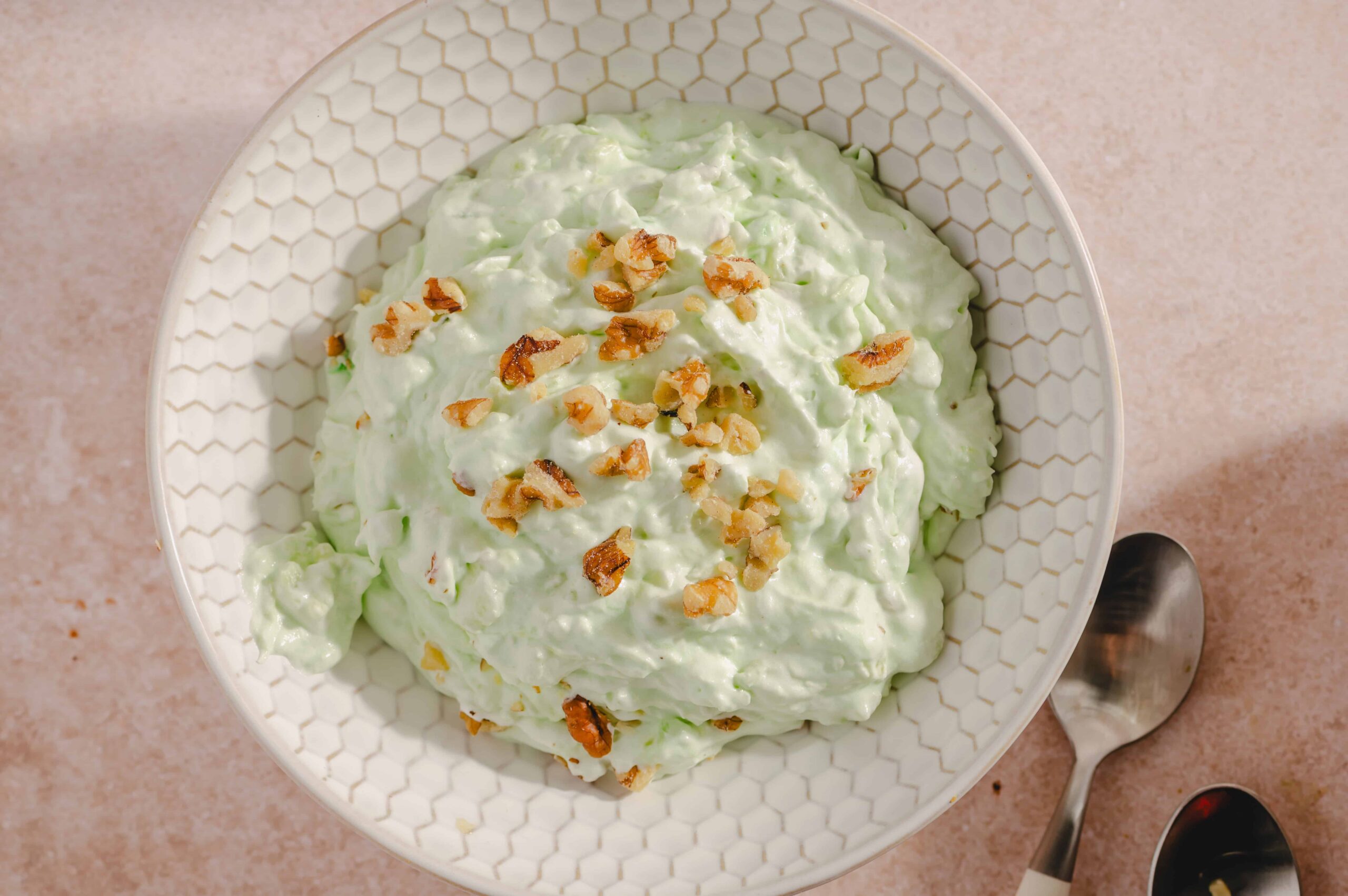 Creamy avocado salad topped with crushed nuts in a white honeycomb-patterned bowl on a light pink surface.
