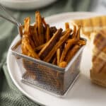 Crispy seasoned sweet potato fries served in a mini wire basket on a white plate with grilled sandwich halves in the background, ideal for enjoyably healthy snack or meal.