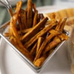 Crispy sweet potato fries in a stainless steel basket with a golden-brown exterior, served with a side of sweet potato wedges in the background, perfect for healthy snack recipes.