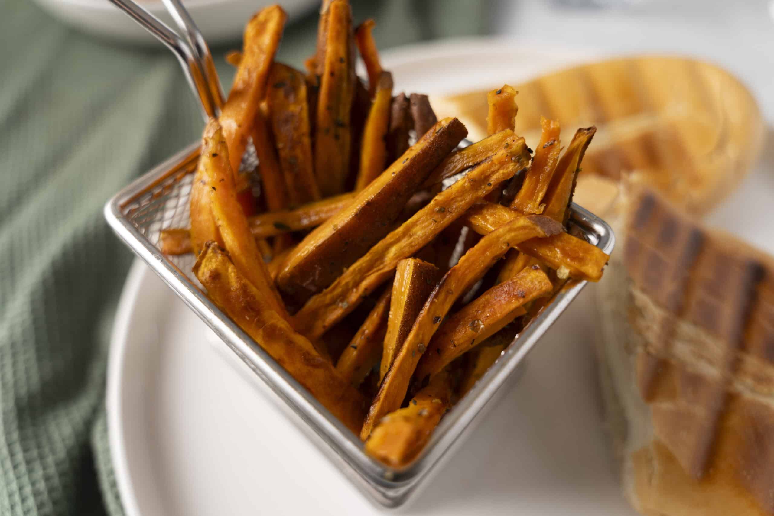 Crispy sweet potato fries in a stainless steel basket with a golden-brown exterior, served with a side of sweet potato wedges in the background, perfect for healthy snack recipes.