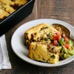 Baked Mexican tamale pie served on a white plate with fresh salsa and guacamole, with the rest of the dish still in a black baking dish on a rustic wooden table.