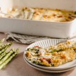 Creamy baked asparagus casserole with golden cheese crust served on a white plate, fresh asparagus stalks beside it, baked in a white ceramic dish, perfect for spring dinners and vegetable lovers.