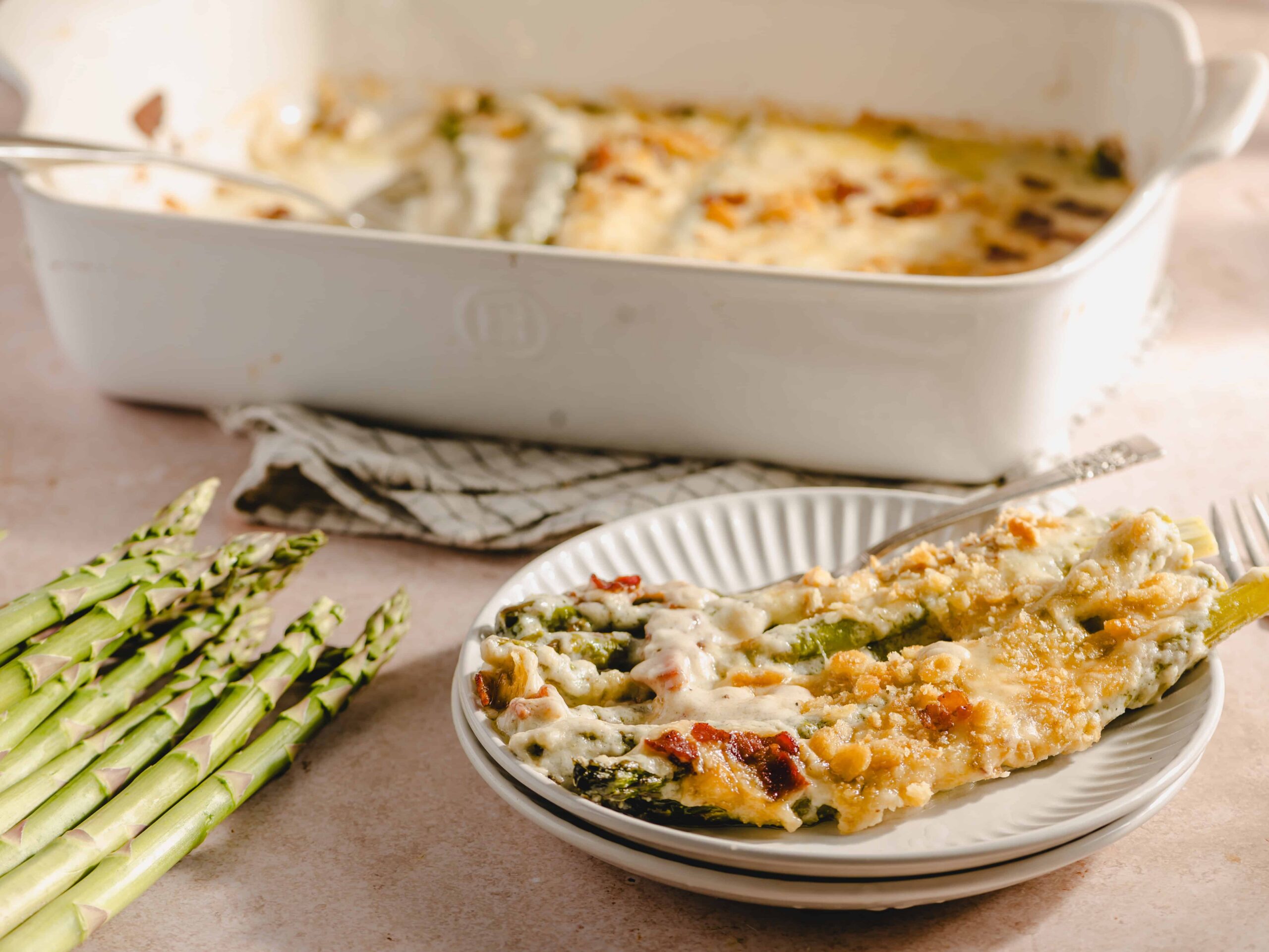 Creamy baked asparagus casserole with golden cheese crust served on a white plate, fresh asparagus stalks beside it, baked in a white ceramic dish, perfect for spring dinners and vegetable lovers.