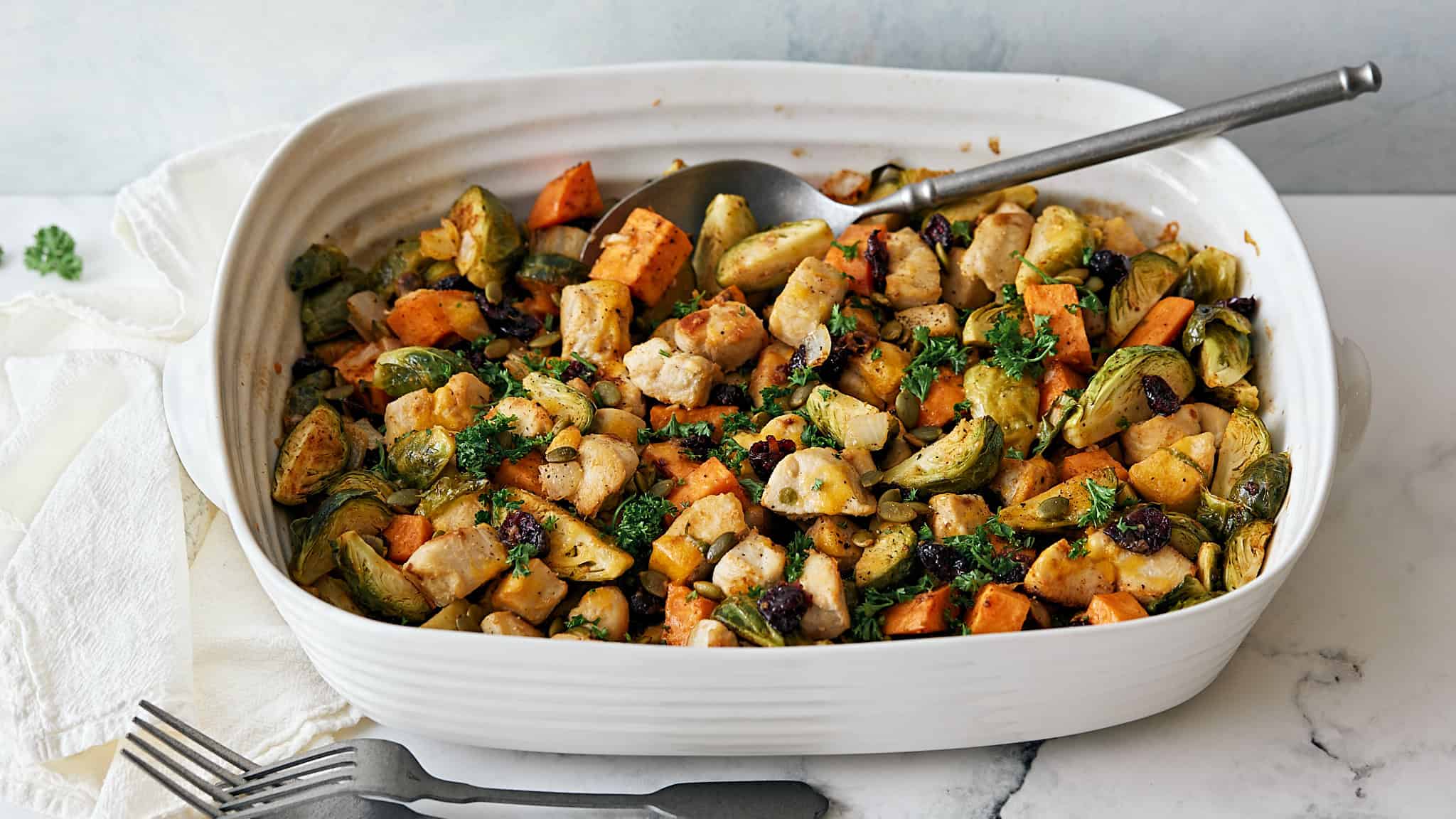 Roasted vegetable medley with Brussels sprouts, sweet potatoes, carrots, and cauliflower in a white ceramic baking dish. Fresh parsley garnish adds color and flavor.