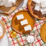 Cream cheese cubes on wooden and ceramic plates, with shredded cheese and a golden spoon, on a checkered tablecloth, perfect for cheese recipes and gourmet food presentation.