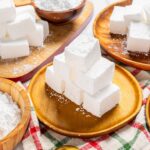 Fresh white sugar cubes on rustic wooden plates and bowls, ready for baking or sweetening beverages, set on a colorful checkered cloth for a cozy kitchen scene.