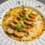 Creamy chicken curry with herbs and spices served on a white decorative plate, garnished with fresh cilantro and green onions.