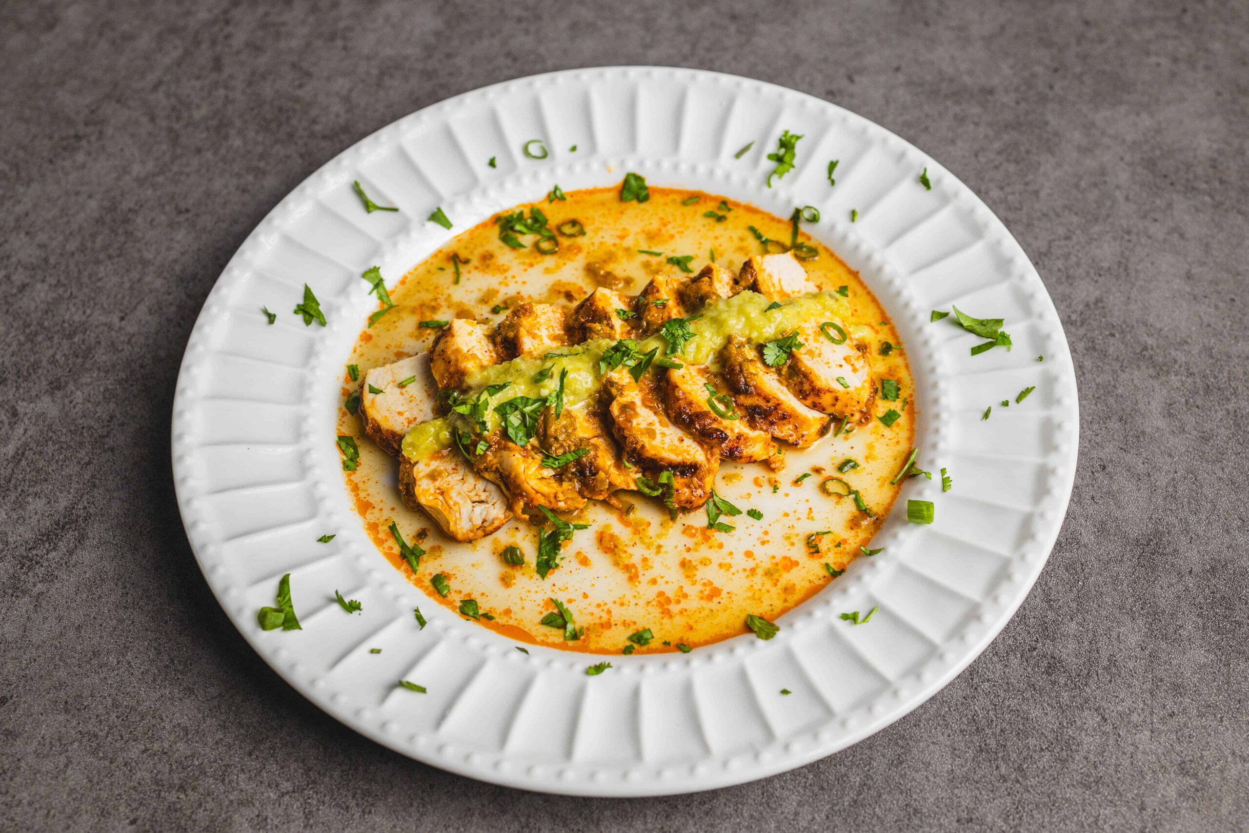Tender grilled chicken slices topped with creamy curry sauce, garnished with fresh cilantro and green onions on a white plate. Perfect for a flavorful dinner or lunch.