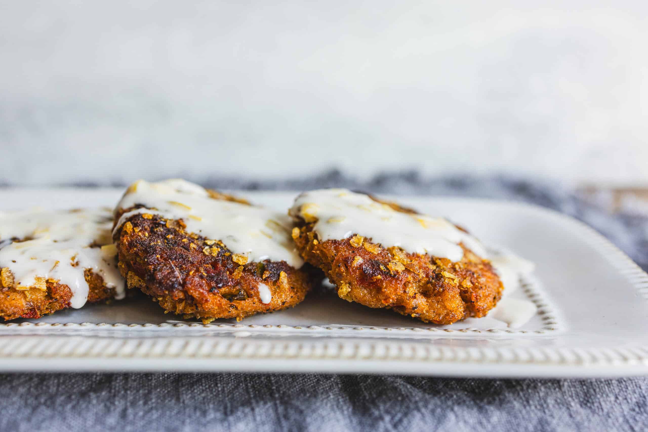 Crispy fried chicken cutlets topped with creamy white sauce, served on an elegant white plate with a dark textured background. Perfect for comfort food recipes and meal ideas.