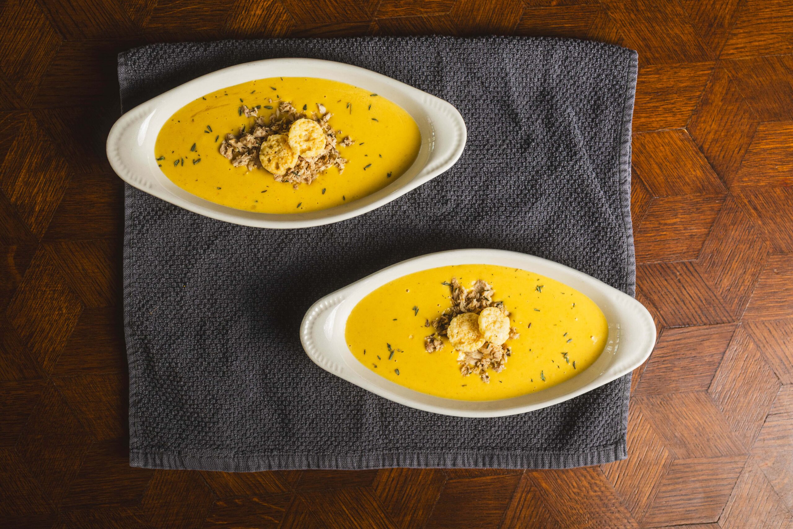 Creamy yellow soup garnished with bread crumbs and herbs served in white oval bowls on a dark cloth and wooden tabletop.