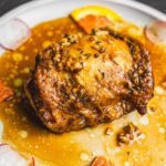 Savory roasted chicken thigh topped with garlic and herbs, served with citrus slices and drizzled with flavorful sauce, on a white plate.