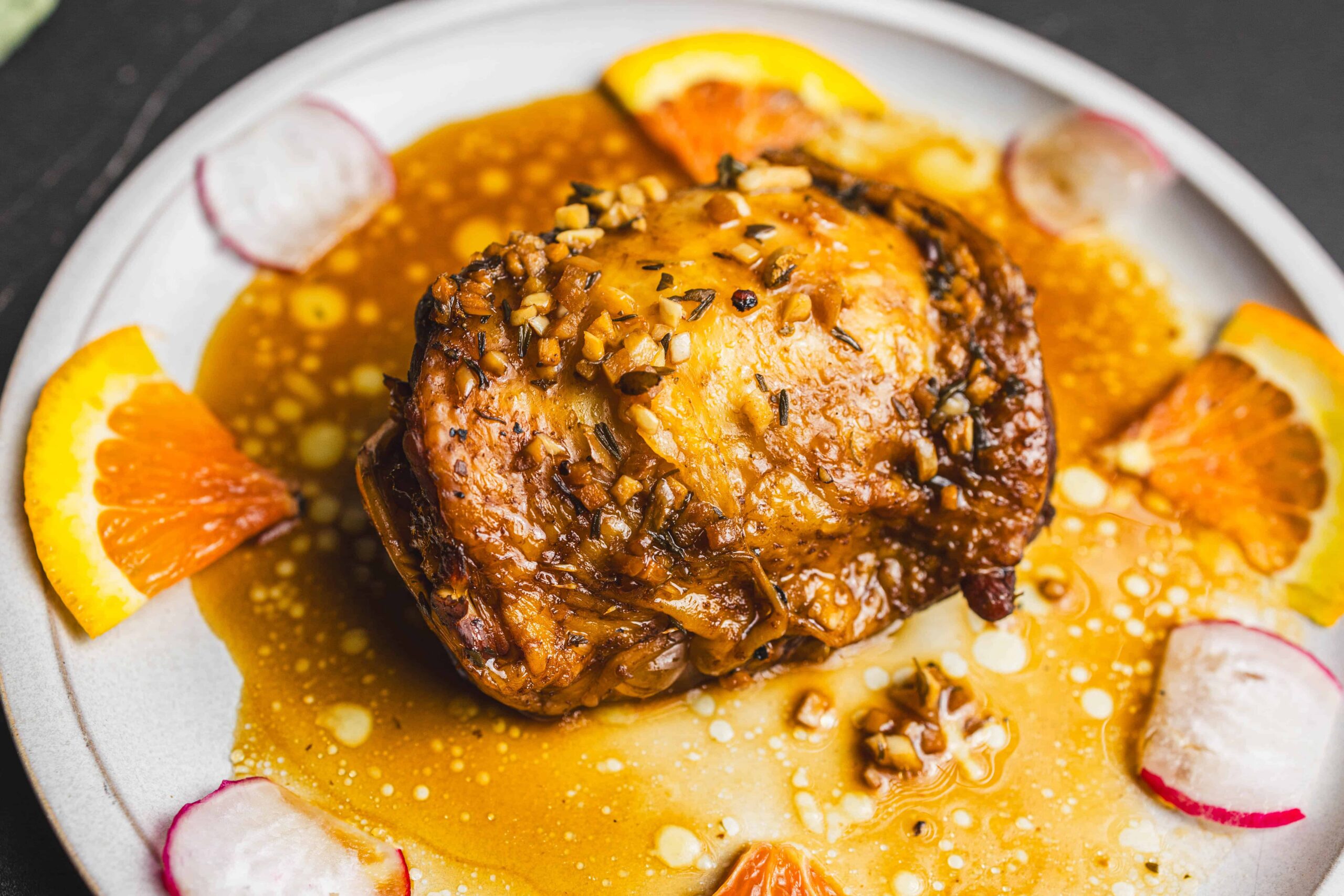 Savory roasted chicken thigh topped with garlic and herbs, served with citrus slices and drizzled with flavorful sauce, on a white plate.