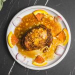 Roasted chicken with orange glaze and crunchy almond topping served with citrus slices and radish garnishes on a white plate. Perfect for flavorful dinner recipes and elegant plating.