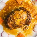 Tender roasted chicken thigh topped with chopped nuts, served on a plate with a creamy orange sauce, garnished with orange slices and radish slices for a flavorful and visually appealing dish.