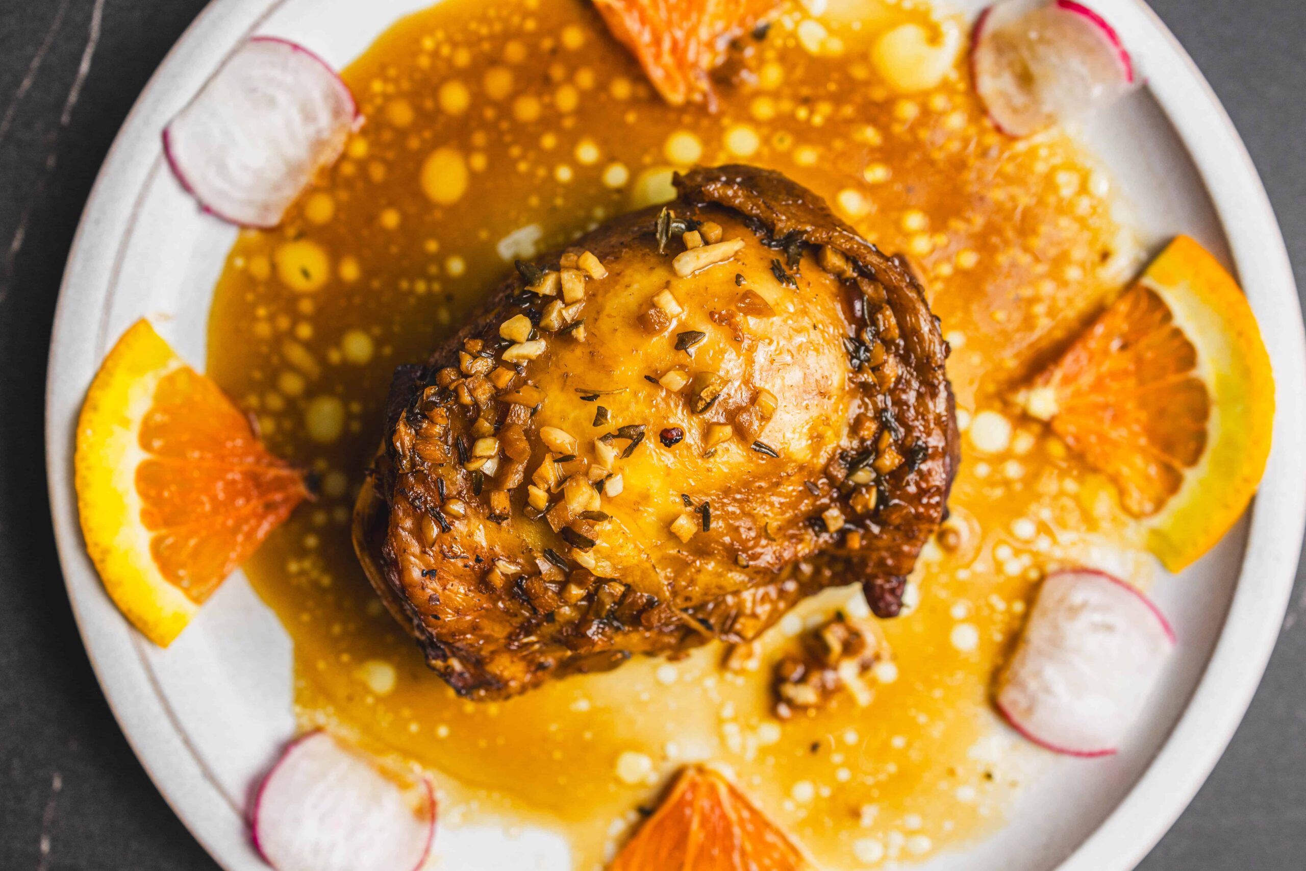 Tender roasted chicken thigh topped with chopped nuts, served on a plate with a creamy orange sauce, garnished with orange slices and radish slices for a flavorful and visually appealing dish.