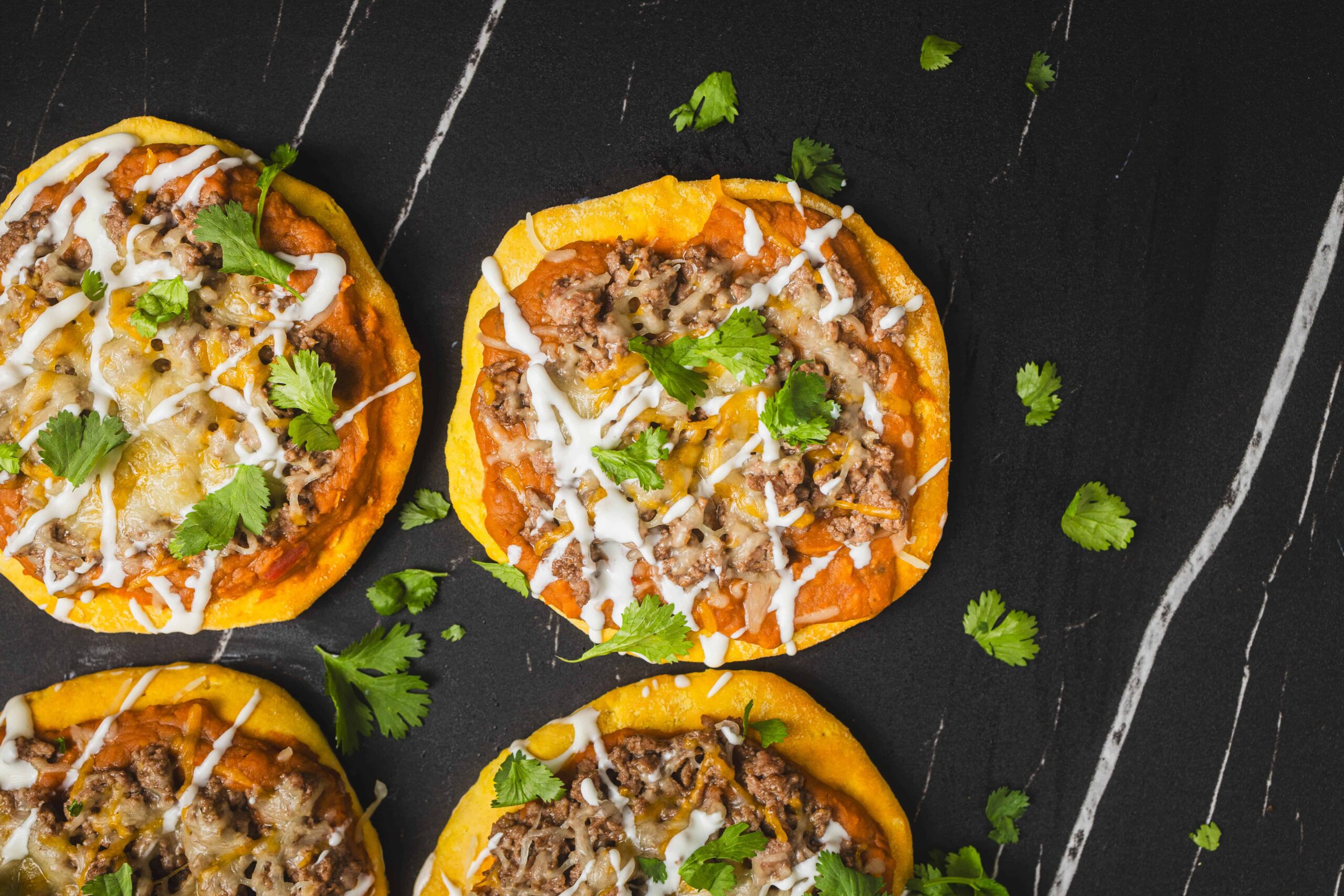 Flavorful mini taco pizzas topped with seasoned ground beef, melted cheese, fresh cilantro, and drizzled with sauce, served on a dark rustic surface for an easy and delicious appetizer or snack.