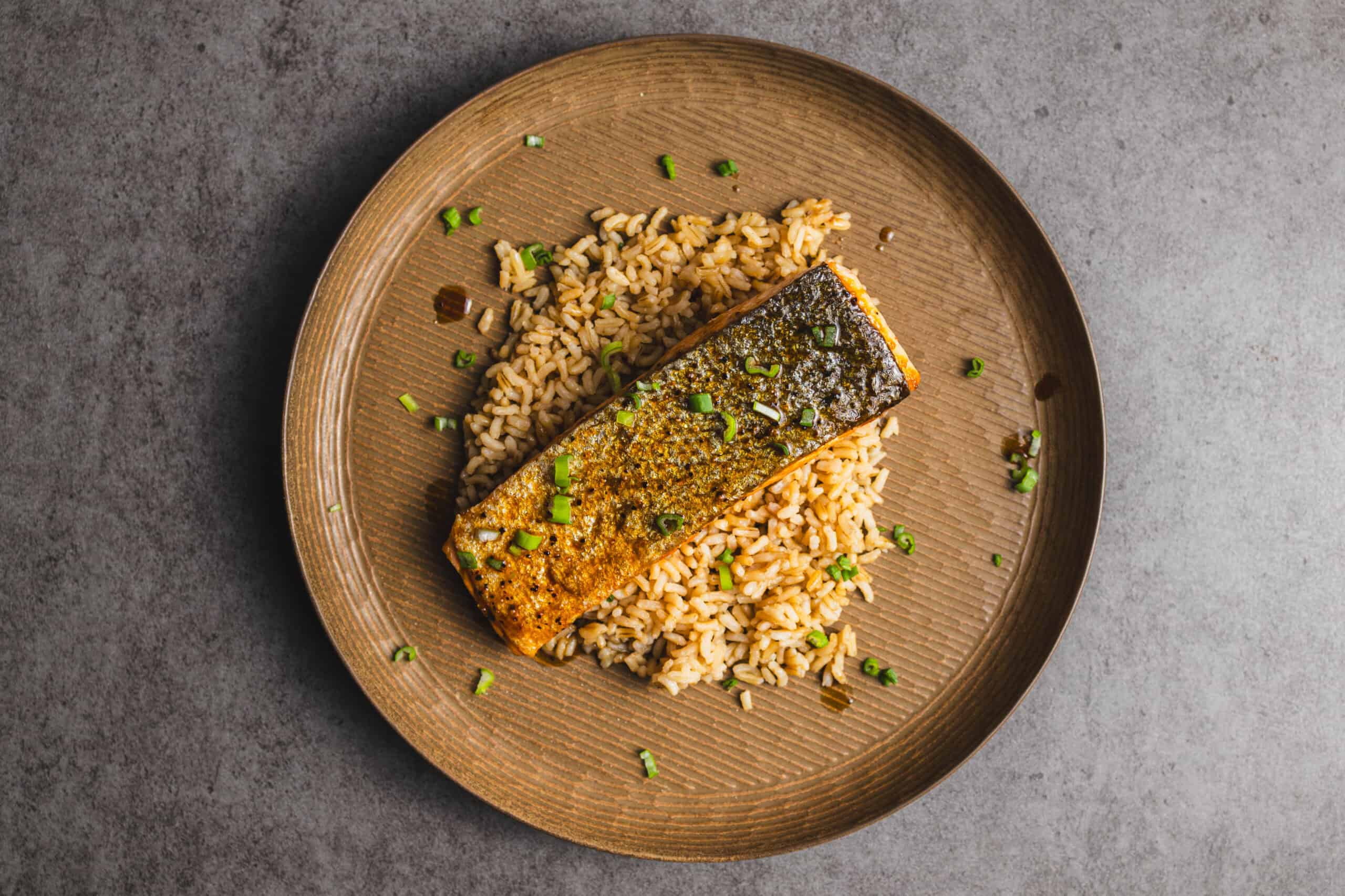 Juicy pan-seared salmon fillet with crispy skin served over seasoned rice and garnished with chopped green onions on a rustic brown plate.