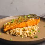 Grilled salmon fillet with crispy skin served over seasoned rice and garnished with chopped green onions on a brown textured plate.