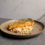 Salmon fillet with crispy skin served over rice on a beige plate with chopped green onions, minimalistic setting, healthy seafood dish, professional food photography.
