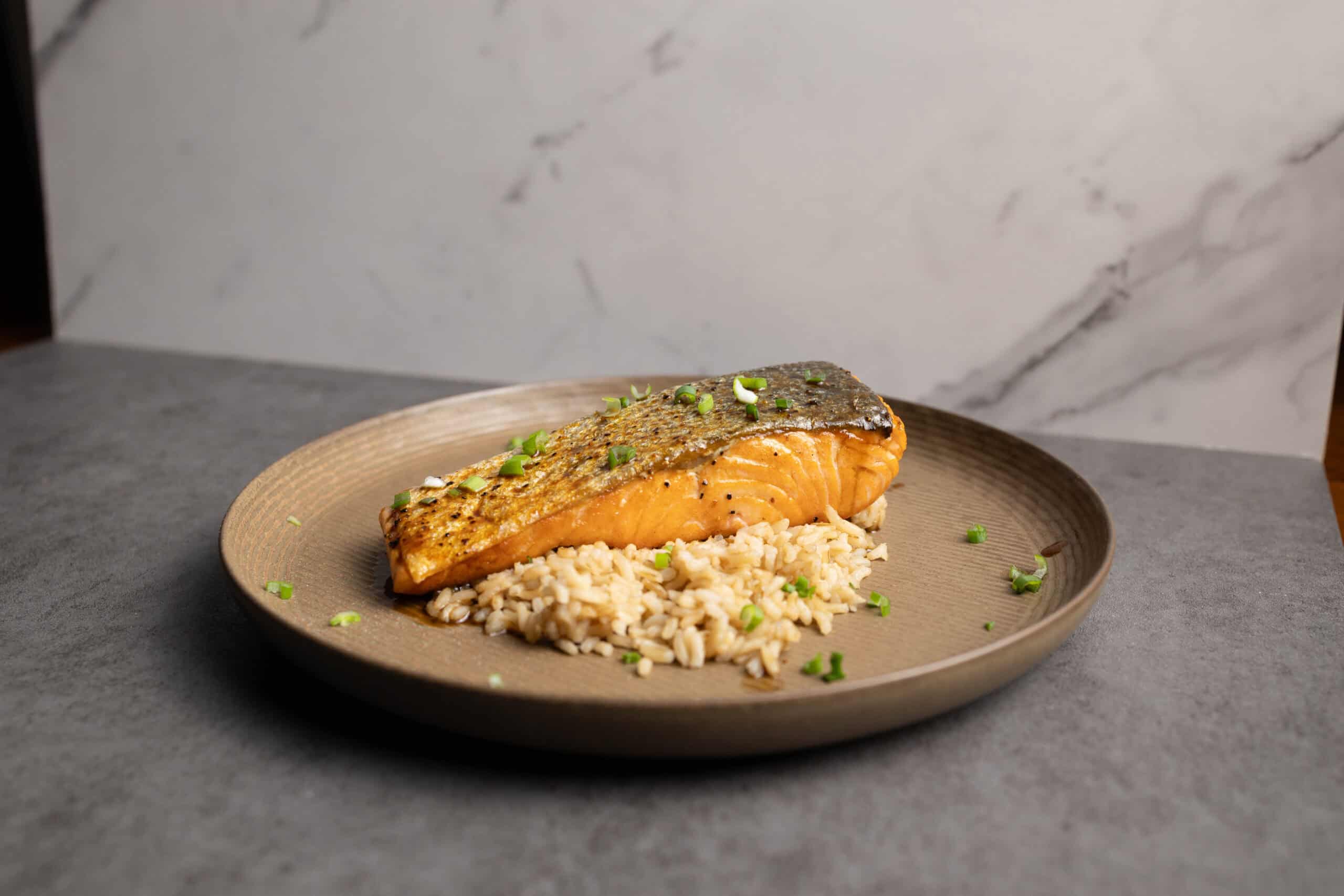 Salmon fillet with crispy skin served over rice on a beige plate with chopped green onions, minimalistic setting, healthy seafood dish, professional food photography.