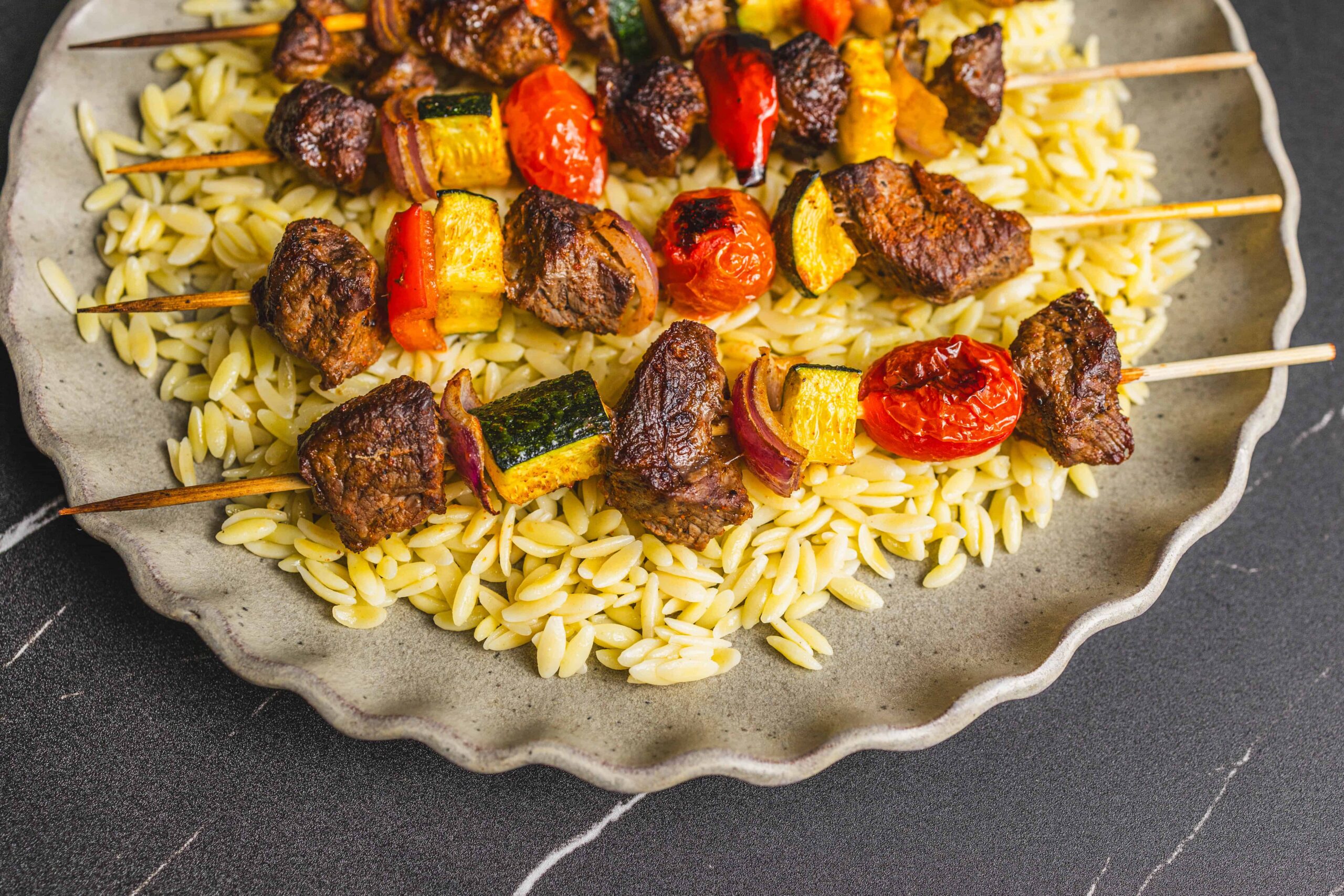 Juicy beef and vegetable skewers on a bed of orzo pasta, served on a rustic gray plate, showcasing a grilled meat and veggie dish perfect for summer barbecues or family dinners.