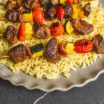Skewered beef and vegetable kebabs served on a bed of orzo pasta on a rustic ceramic plate. The skewers feature grilled beef chunks, zucchini, cherry tomatoes, red onions, and yellow bell peppers, perfect for BBQ or summer grilling.