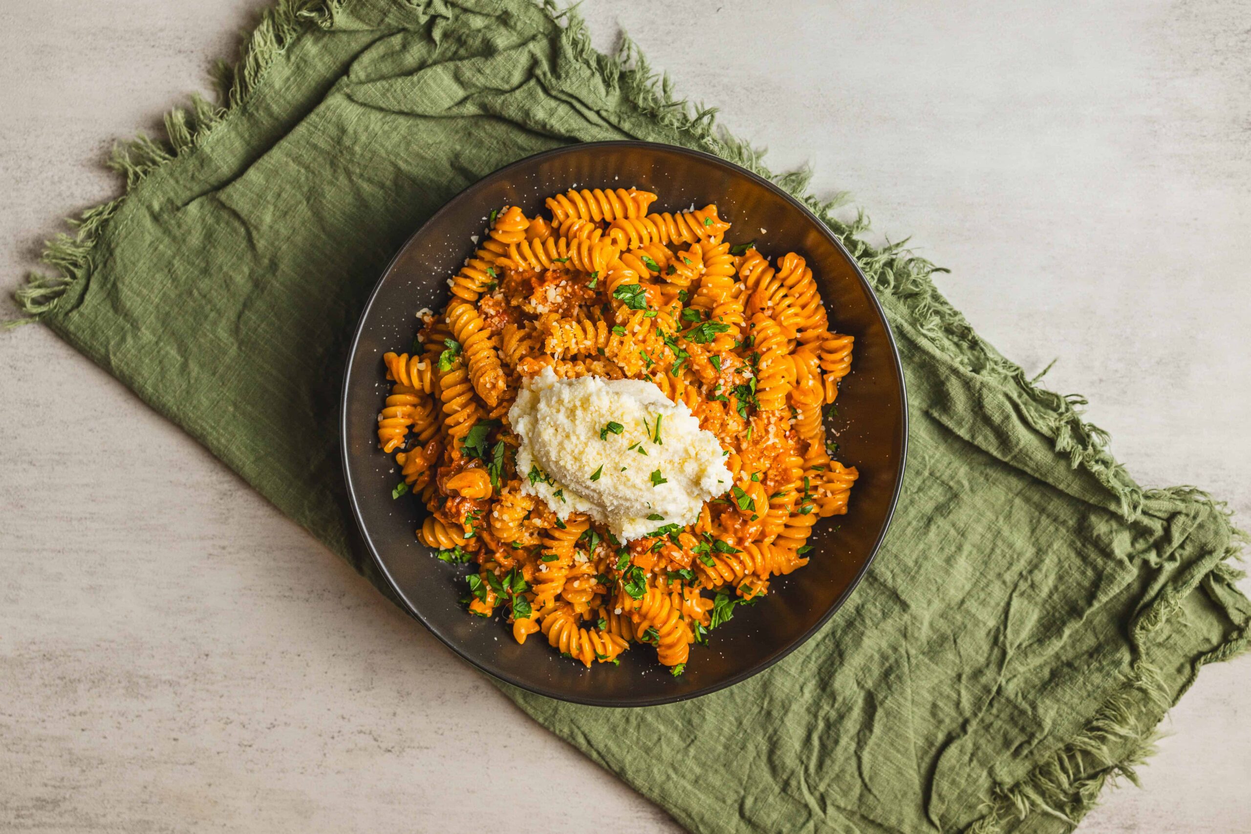 Creamy tomato sauce fusilli pasta topped with grated cheese and fresh herbs in a black bowl on a rustic green cloth.