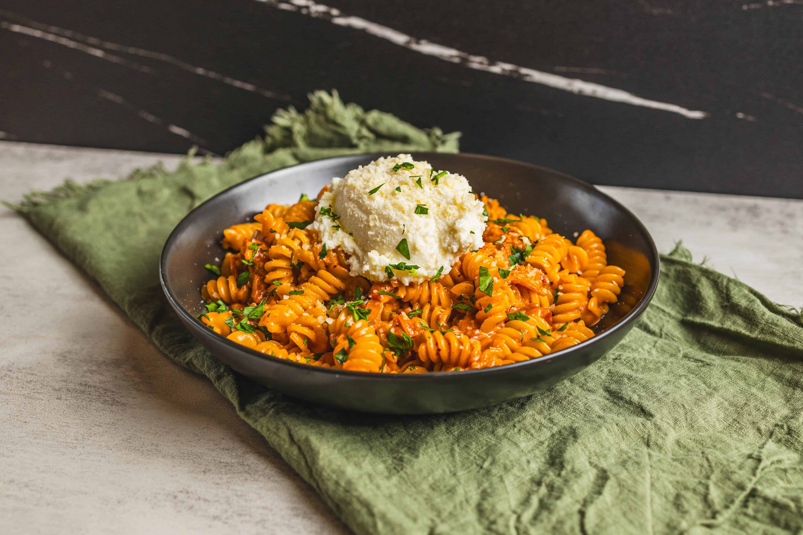 Creamy tomato pasta fusilli with basil and a dollop of ricotta cheese on top, served in a black bowl on a rustic table with green cloth. Perfect comfort food for pasta lovers.