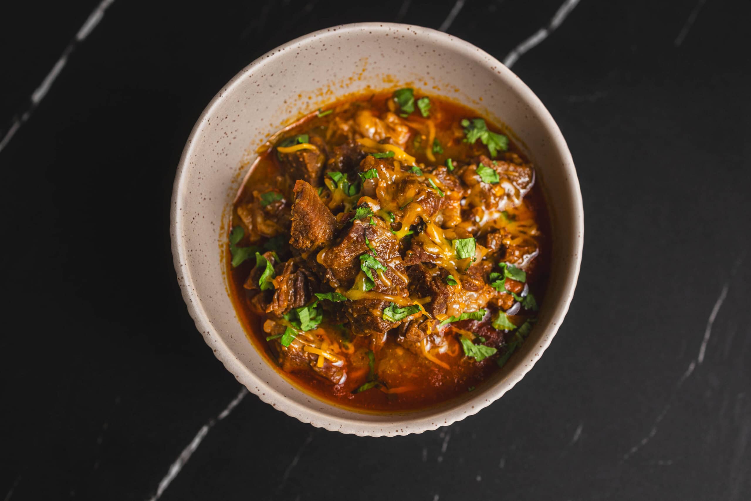 Savory beef chili in a white bowl garnished with fresh cilantro on a dark marble surface.