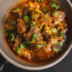 Tender beef chili with melted cheese and chopped cilantro in a beige bowl on a dark background, showcasing a flavorful and hearty comfort food dish.