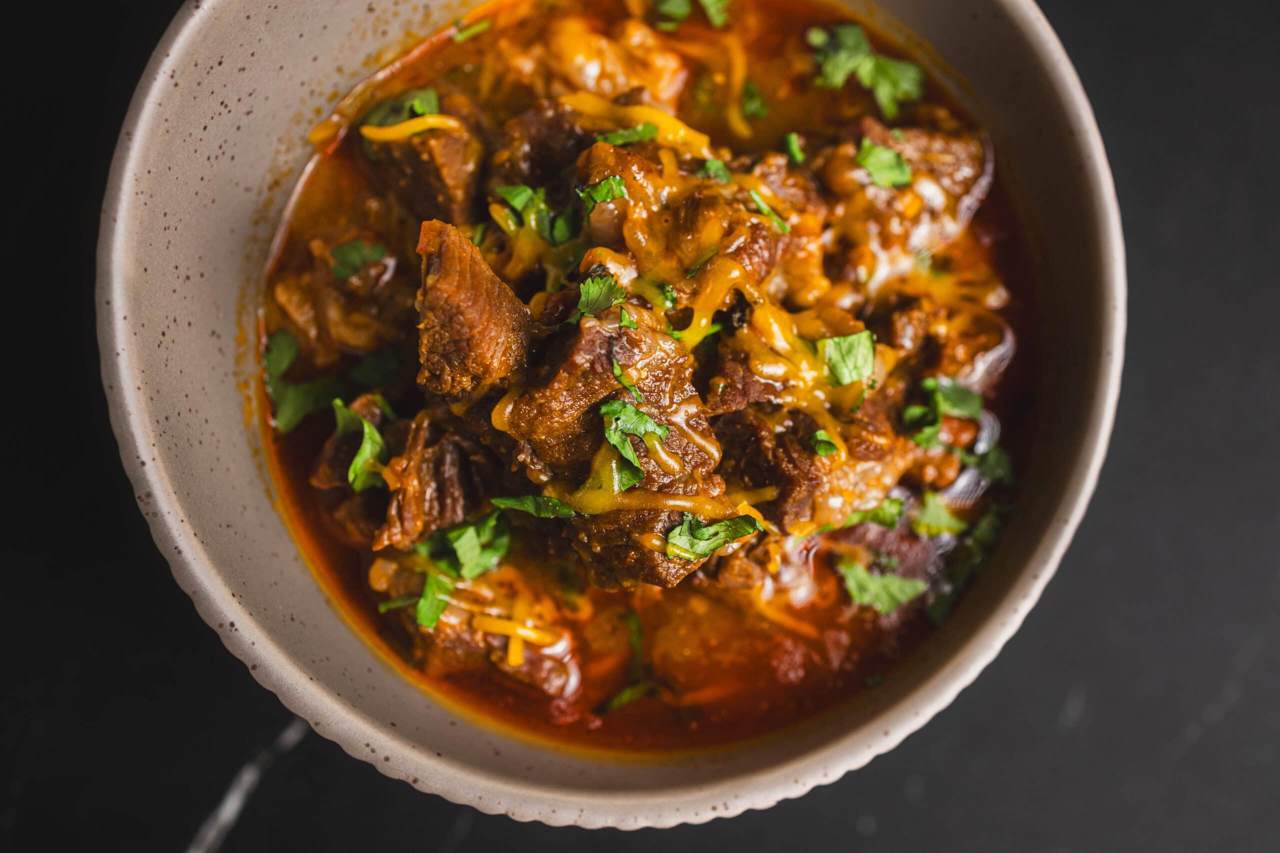 Tender beef chili with melted cheese and chopped cilantro in a beige bowl on a dark background, showcasing a flavorful and hearty comfort food dish.