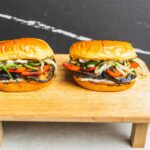 Grilled eggplant and vegetable sandwiches on toasted buns served on a wooden cutting board with a black background.