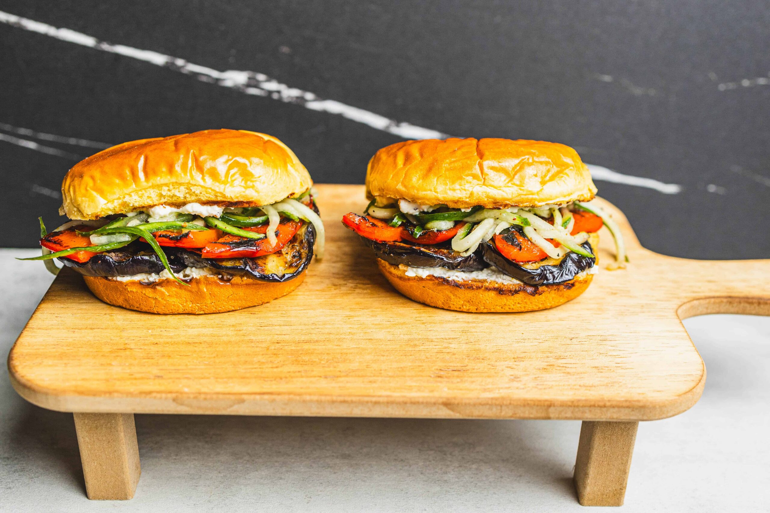 Grilled eggplant and vegetable sandwiches on toasted buns served on a wooden cutting board with a black background.