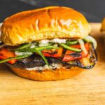 Grilled veggie burger with roasted peppers, grilled eggplant, fresh cucumbers, and herbs on a golden bun on a wooden board.