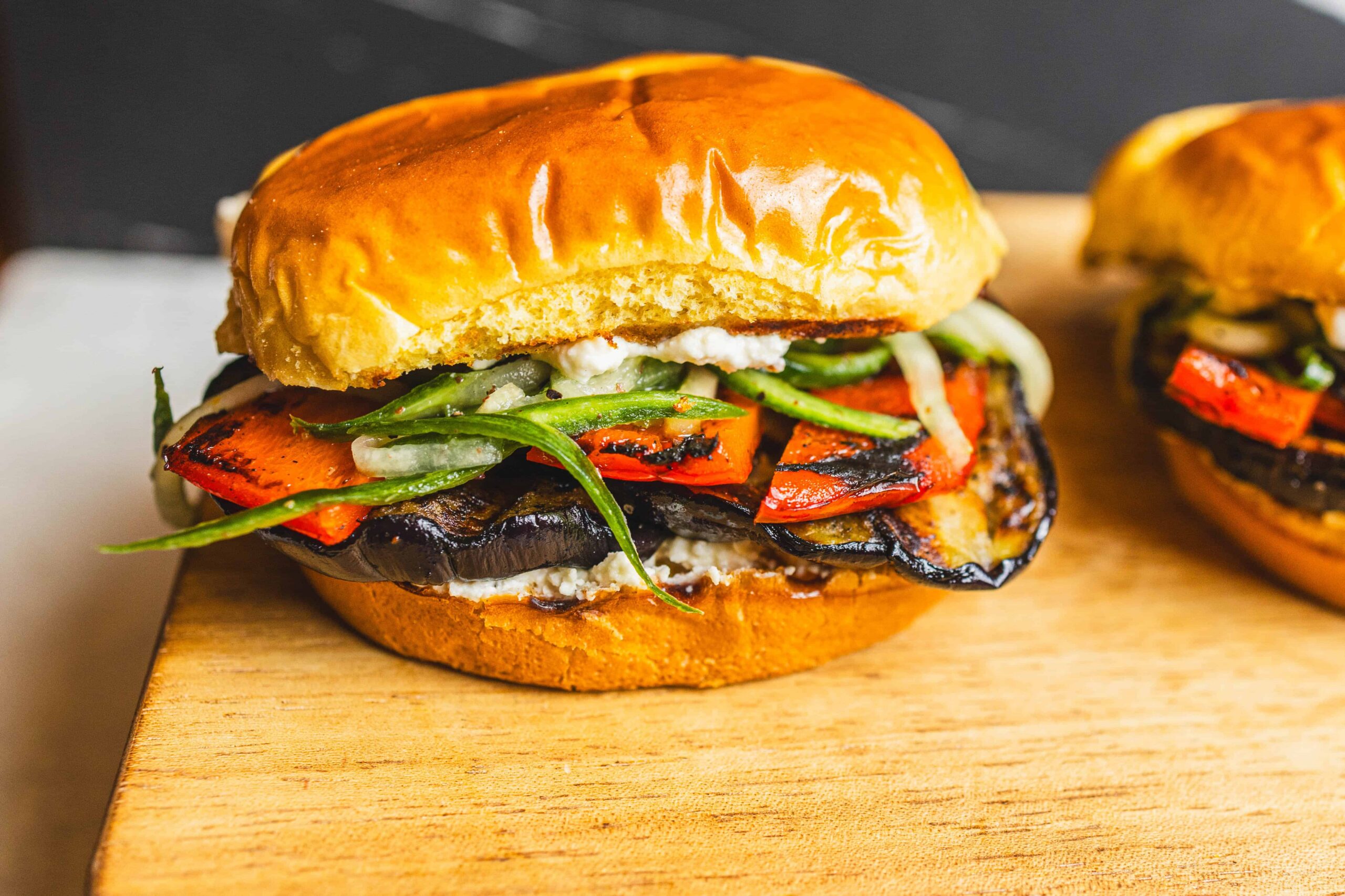 Grilled veggie burger with roasted peppers, grilled eggplant, fresh cucumbers, and herbs on a golden bun on a wooden board.